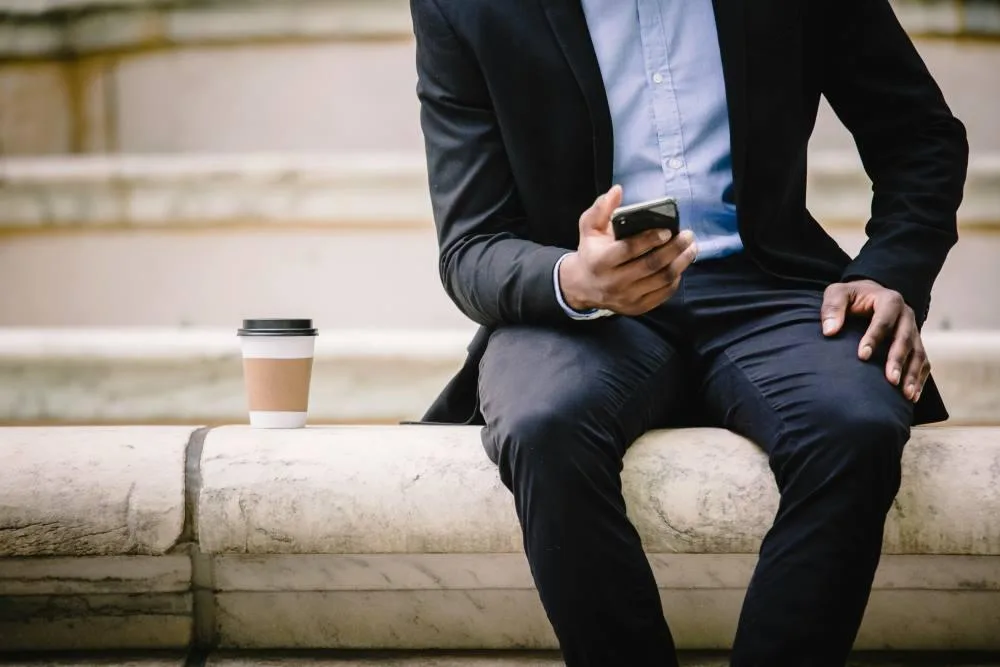 Crop businessman using smartphone while resting on bench with takeaway coffee